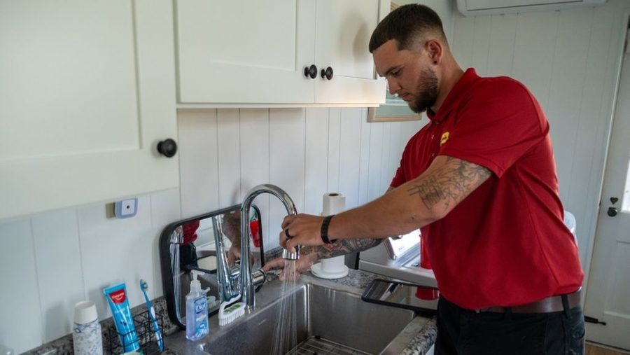 Technician performing drain service by inspecting and testing a kitchen sink faucet during a residential plumbing service call.