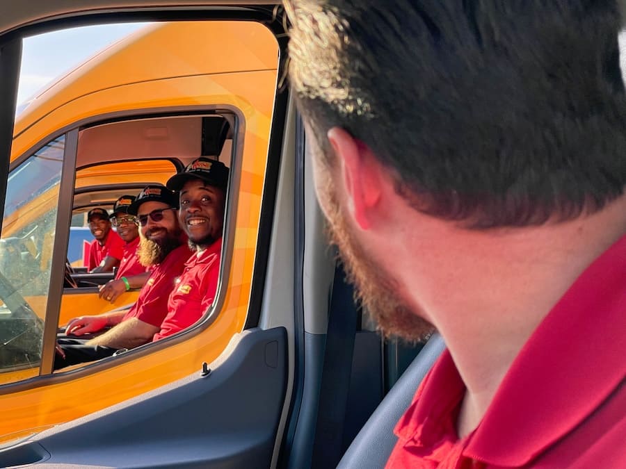 People wearing red shirts sitting in a yellow delivery truck cab, smiling at the camera; foreground person in red shirt in the passenger seat observes.