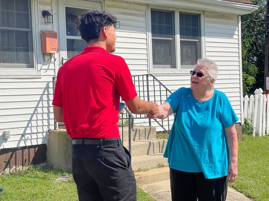 Man in a red polo shakes hands with an elderly woman in a turquoise shirt outside a white house, smiling at each other.
