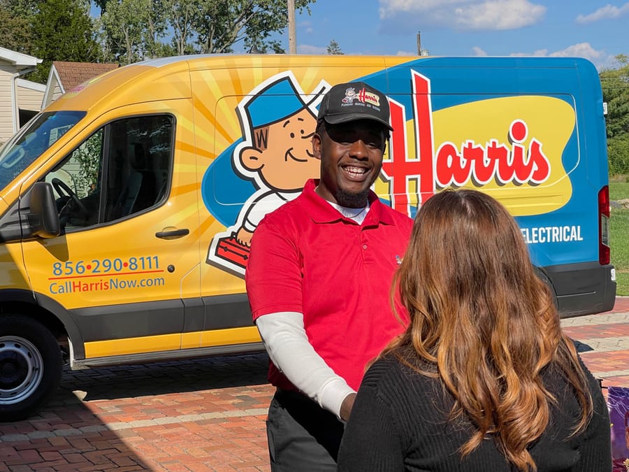 A Harris technician in a red uniform greets a homeowner beside a bright yellow service van, highlighting professional and friendly water heater service.