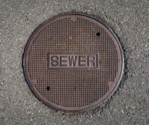 Round metal sewer manhole cover with a grid pattern and a recessed 'SEWER' label, set in asphalt pavement.