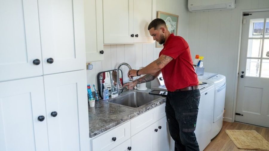 Professional plumber performing plumbing repairs beneath a kitchen sink, inspecting and servicing the faucet and plumbing connections in a residential home.